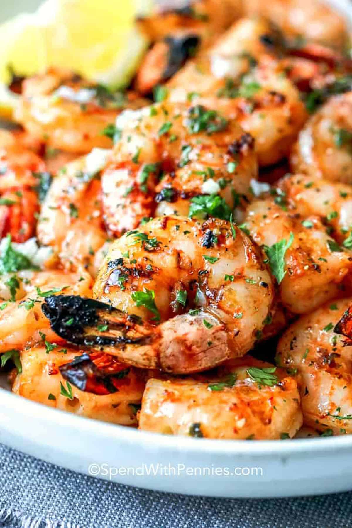 Close-up of grilled shrimp garnished with chopped parsley and spices, served on a white plate with a wedge of lemon in the background.