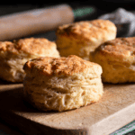 Four golden, flaky sourdough biscuits sit on a wooden cutting board with a rolling pin and a bowl of flour blurred in the background. Warm light highlights the biscuits’ textured, layered edges.