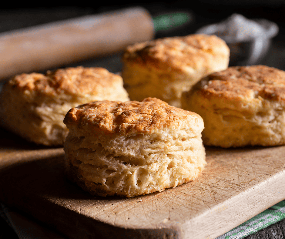 Four golden, flaky sourdough biscuits sit on a wooden cutting board with a rolling pin and a bowl of flour blurred in the background. Warm light highlights the biscuits’ textured, layered edges.