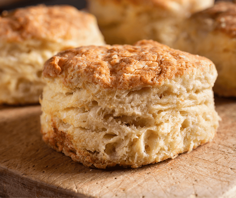 A close-up of a golden, flaky sourdough biscuit sits on a wooden surface, showing its layered texture. Other sourdough biscuits are blurred in the background.