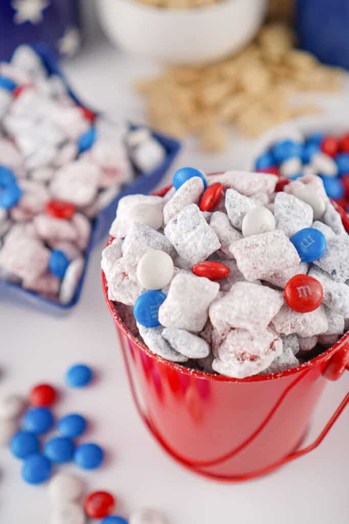 A red bucket filled with 4th of July Puppy Chow—white powdered snack mix and red, white, and blue candy-coated chocolates—with more candy and snack mix scattered in the background.
