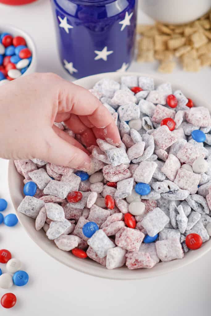 A hand reaches into a bowl of 4th of July Puppy Chow, a patriotic snack mix made with red, white, and blue M&M’s and powdered sugar-coated cereal pieces, with themed decorations and candies in the background.