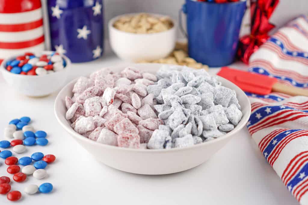 A white bowl filled with 4th of July Puppy Chow sits on a table adorned with patriotic-themed decor, American flag patterns, red, white, and blue candies, and star-printed containers.