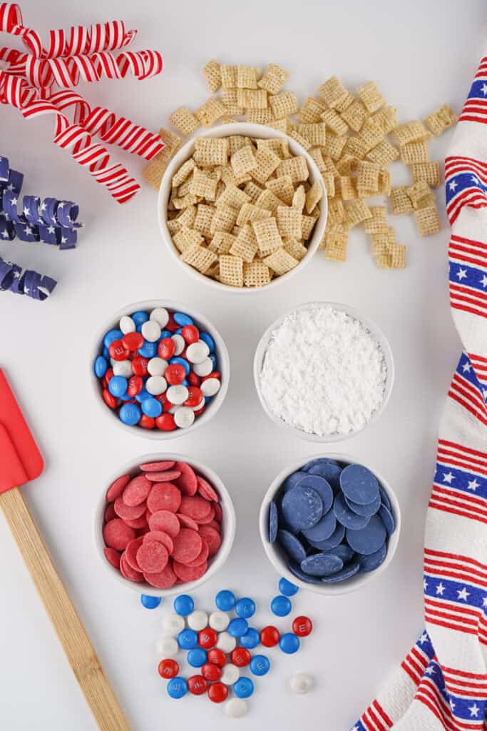Bowls with 4th of July Puppy Chow made from Chex cereal, powdered sugar, red and blue candy melts, and patriotic M&M’s sit on a white surface, surrounded by festive ribbon decorations and a striped cloth.
