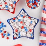 A star-shaped blue bowl filled with 4th of July Puppy Chow and red, white, and blue M&M’s, surrounded by a matching striped patriotic napkin and scattered candies on a white surface.