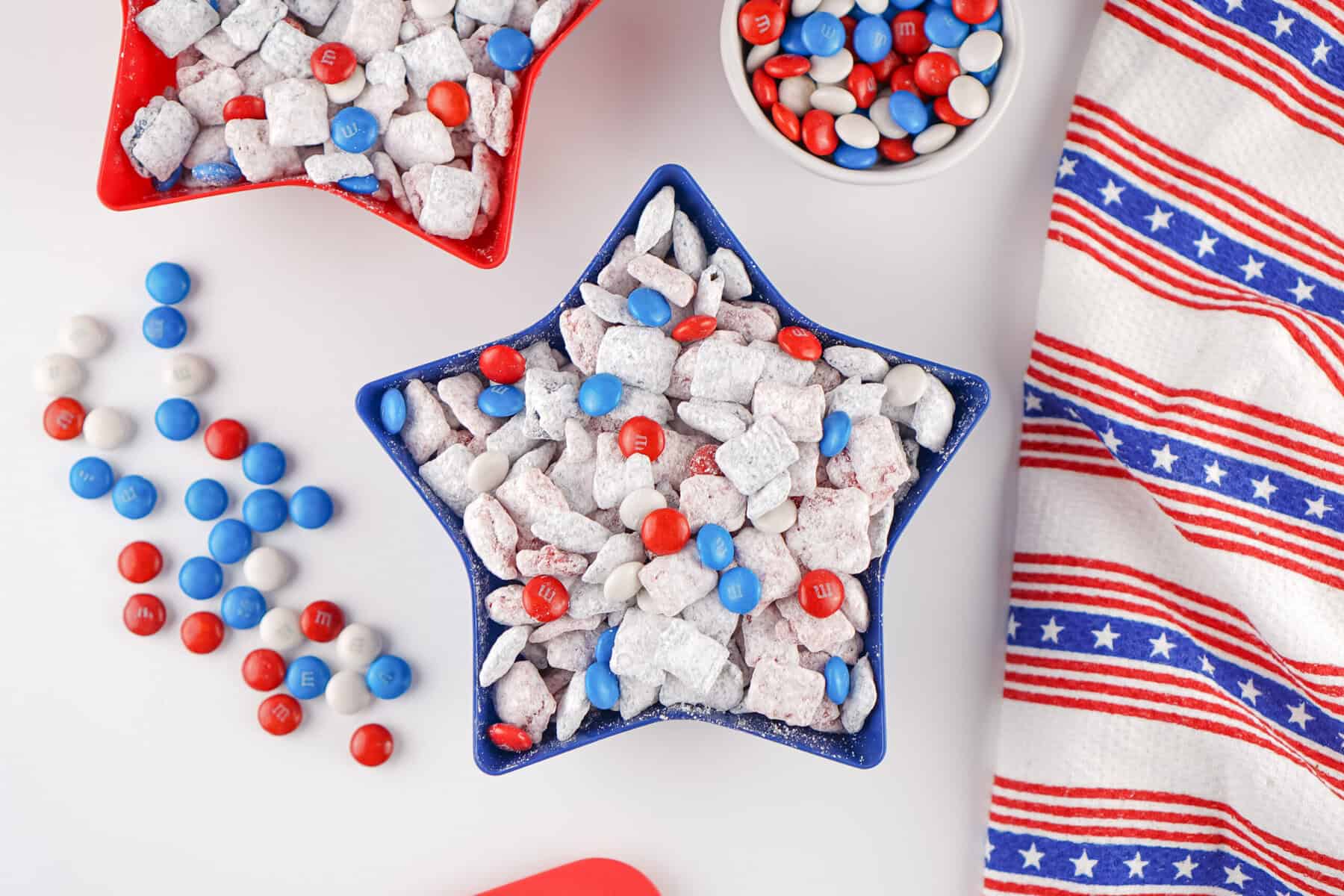 A star-shaped blue bowl filled with 4th of July Puppy Chow and red, white, and blue M&M’s, surrounded by a matching striped patriotic napkin and scattered candies on a white surface.