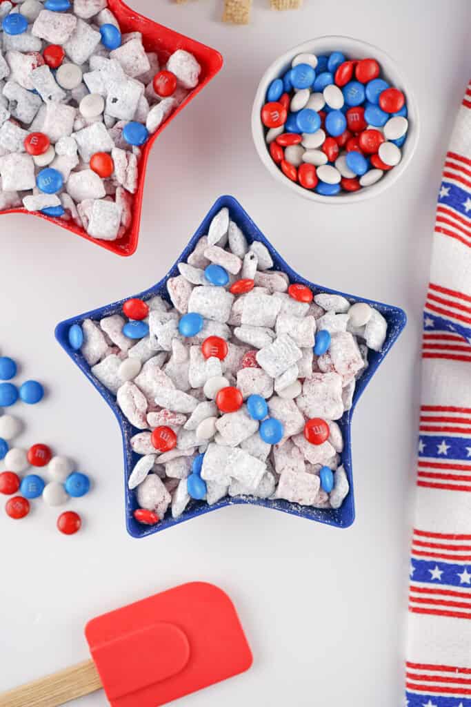A red, white, and blue star-shaped bowl filled with 4th of July Puppy Chow dusted in powdered sugar and sprinkled with festive M&Ms, alongside a matching bowl of M&Ms and a patriotic striped towel.