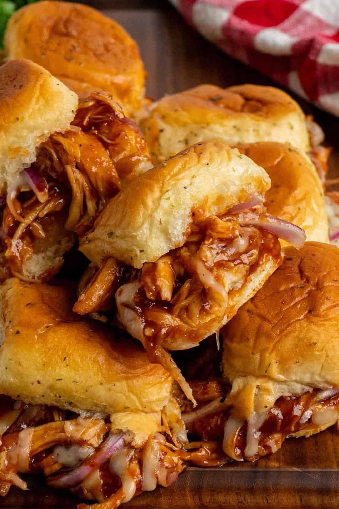 A close-up of several BBQ chicken sliders with shredded chicken, melted cheese, and red onions on soft buns, stacked on a wooden surface. A red and white checkered cloth is visible in the background.
