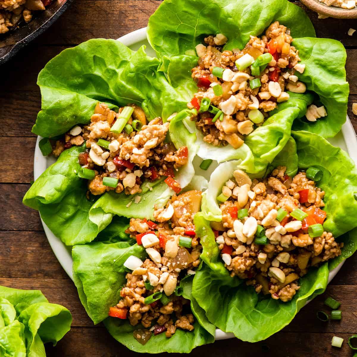 A plate of fresh lettuce leaves topped with a mixture of diced vegetables, ground meat, chopped nuts, and green onions, served as lettuce wraps on a wooden table.