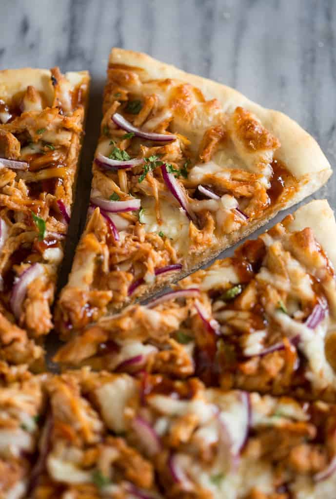 A close-up of a sliced pizza topped with shredded chicken, red onion, melted cheese, and fresh herbs on a thin crust, displayed on a marble surface.