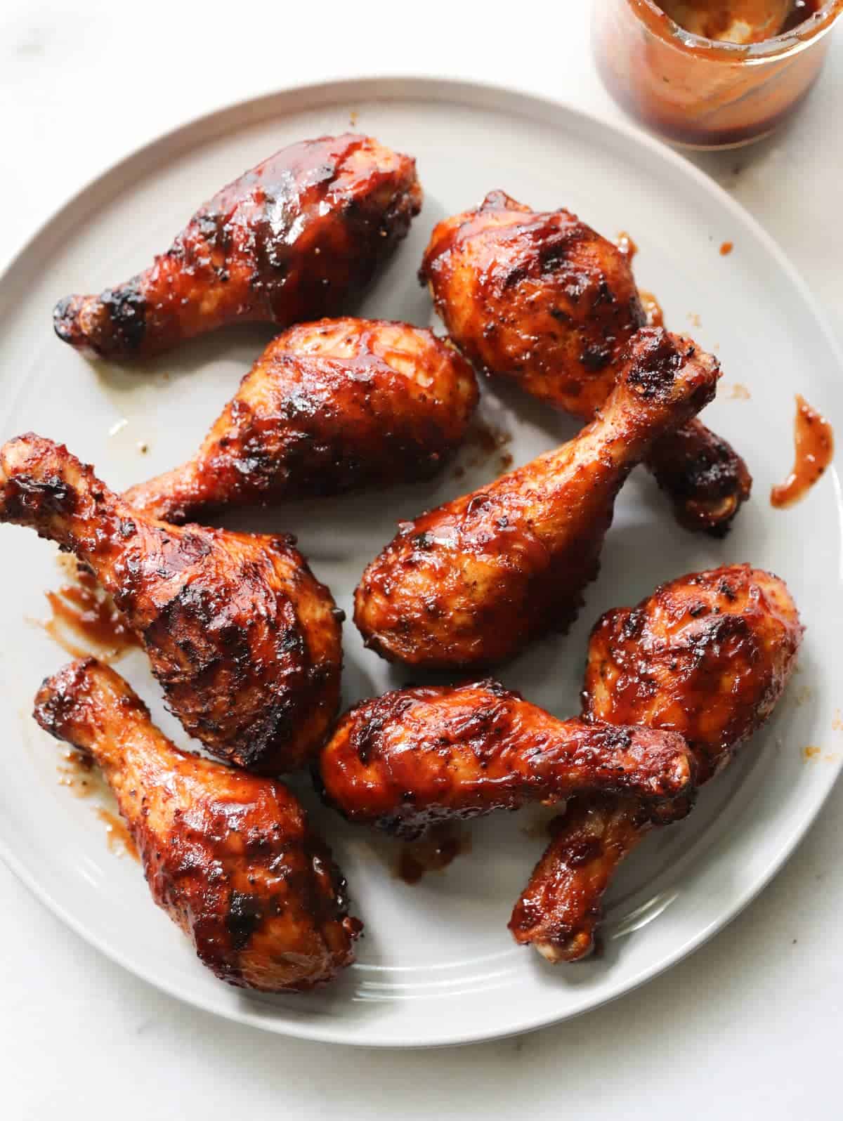 Eight grilled chicken drumsticks coated in a shiny, dark barbecue sauce are arranged on a white plate. A jar with extra sauce is partially visible in the upper right corner.