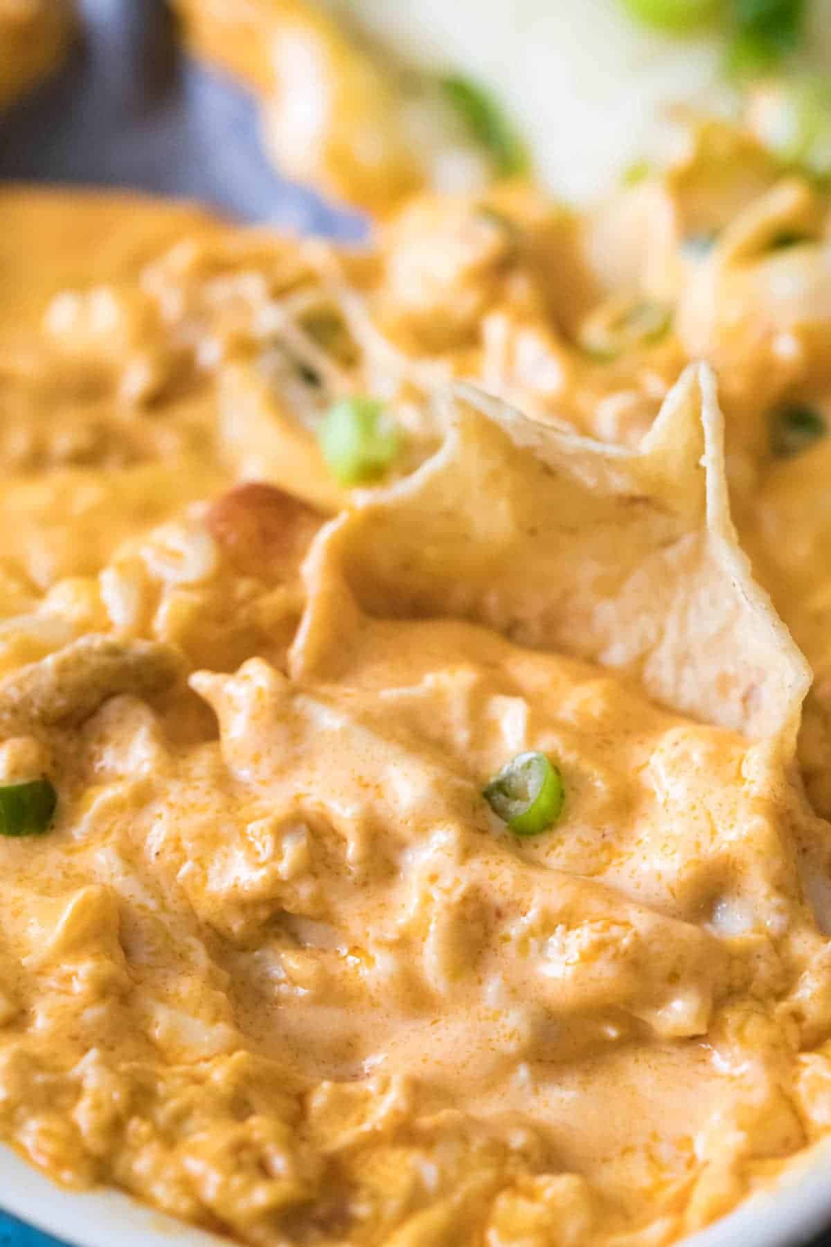 A close-up of a tortilla chip dipped in a creamy, orange-colored cheesy dip, garnished with small pieces of green onion.