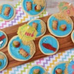 Colorful beach sugar cookies decorated with blue icing, graham cracker sand, gummy fish and bears, mini umbrellas, and candy pearls are arranged on a wooden tray and zigzag-patterned napkin.