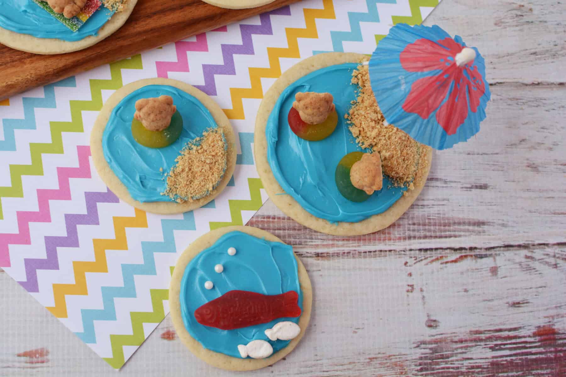 Beach sugar cookies decorated with blue icing, gummy candies, crushed cookie “sand,” and animal crackers resemble a charming beach scene. One cookie features a paper umbrella, another has a red gummy fish and white icing “bubbles.”.