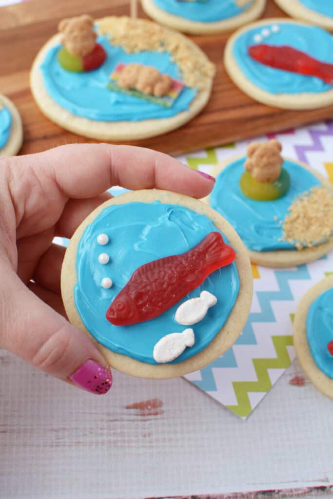 A hand holds a round Beach sugar cookie decorated with blue frosting, white candy bubbles, a red gummy fish, and two small white candy fish. More Beach sugar cookies are on the table in the background.