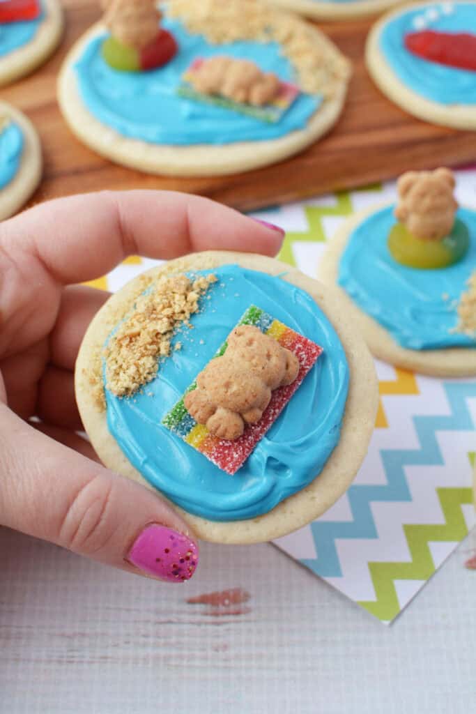 A hand with pink nails holds a beach sugar cookie topped with blue frosting, a gummy candy, a teddy bear-shaped cookie, and cookie crumbs, designed to look like a bear lounging at the beach. More decorated cookies are in the background.