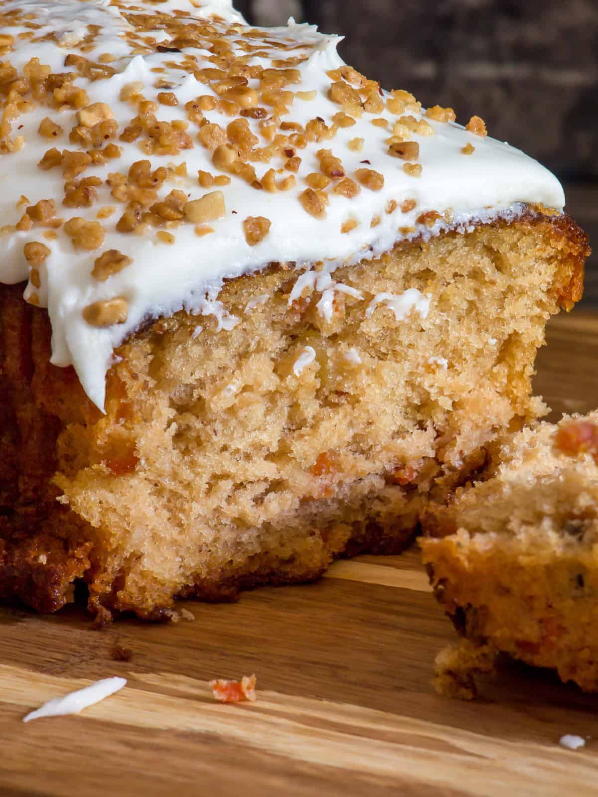 A close-up of a sliced loaf cake topped with white frosting and sprinkled with chopped nuts, displayed on a wooden surface. One slice is partially cut and lies in front of the loaf.