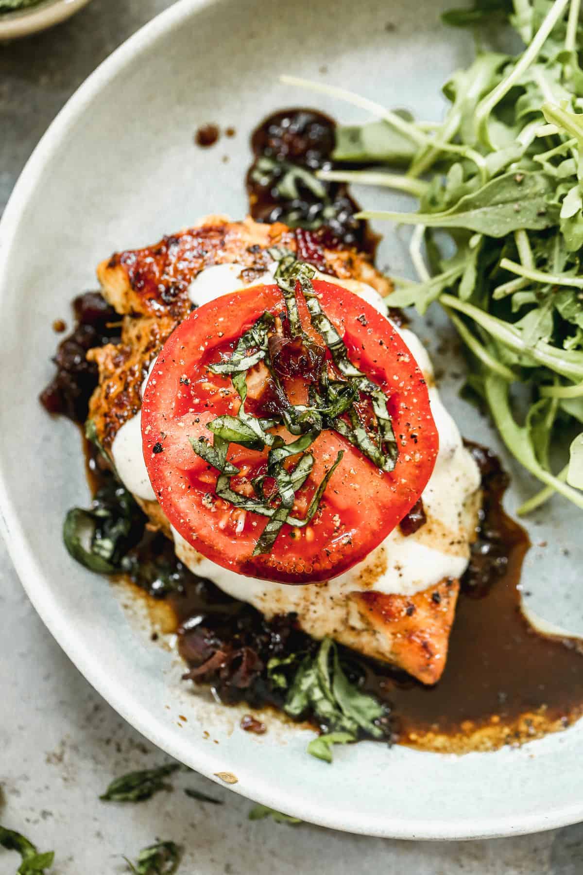 A plate with grilled chicken breast topped with melted mozzarella, a slice of tomato, chopped basil, and balsamic glaze, served next to a small pile of fresh arugula.