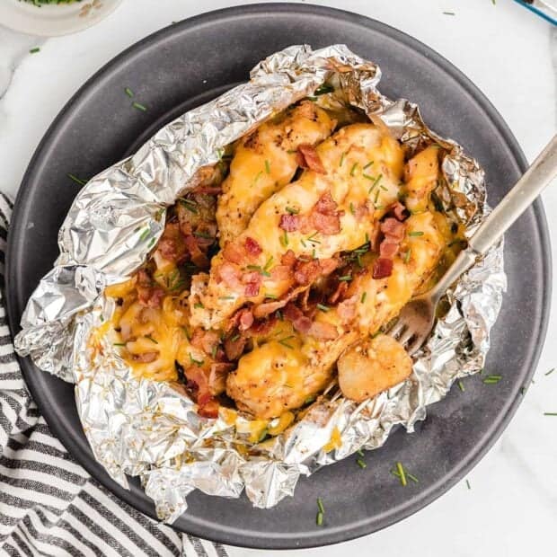 Foil packet filled with cooked chicken, melted cheese, potato slices, and crispy bacon bits, garnished with chopped chives and served on a dark plate with a fork.