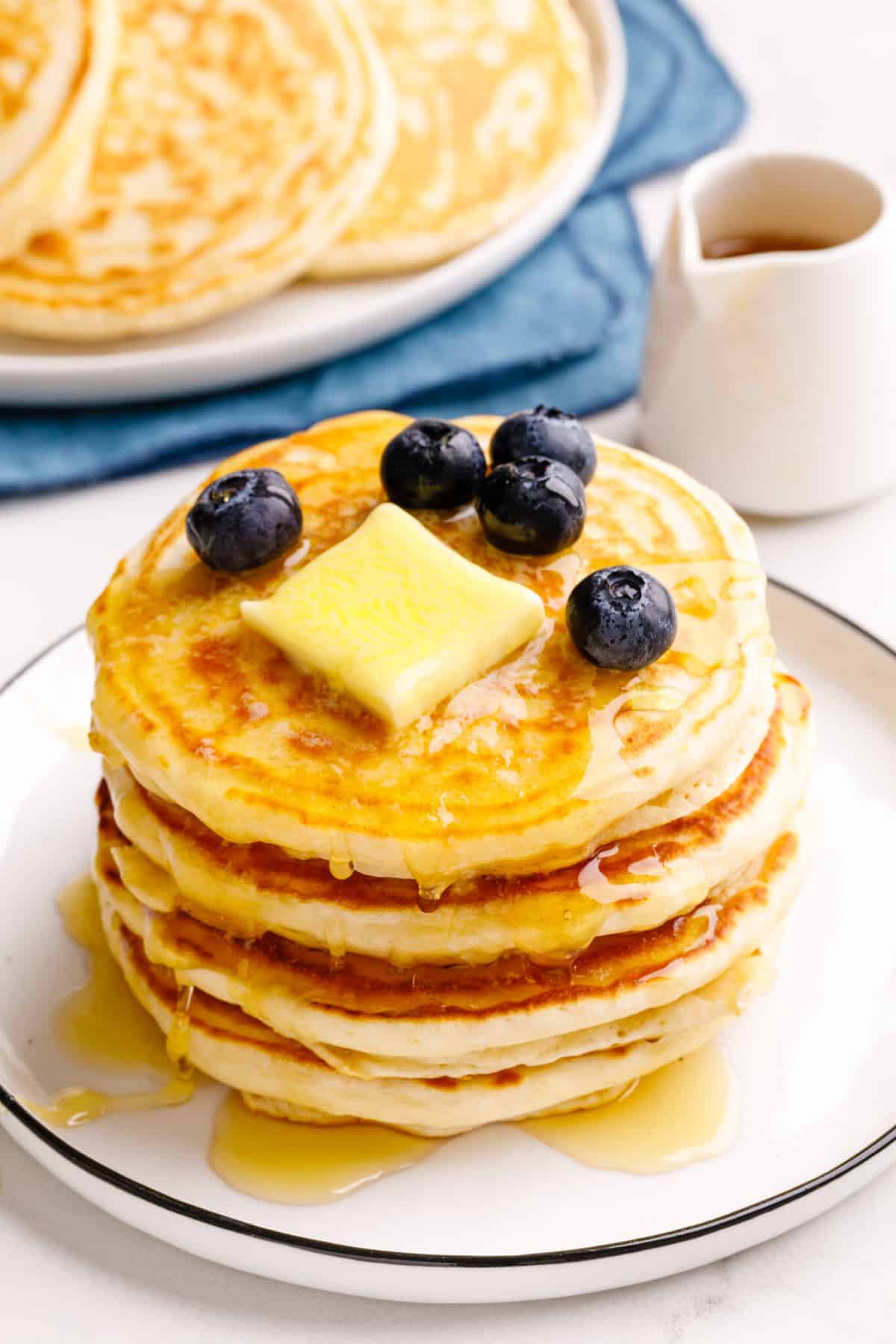A stack of pancakes topped with a pat of butter, fresh blueberries, and syrup on a white plate, with extra pancakes and a small pitcher of syrup in the background.