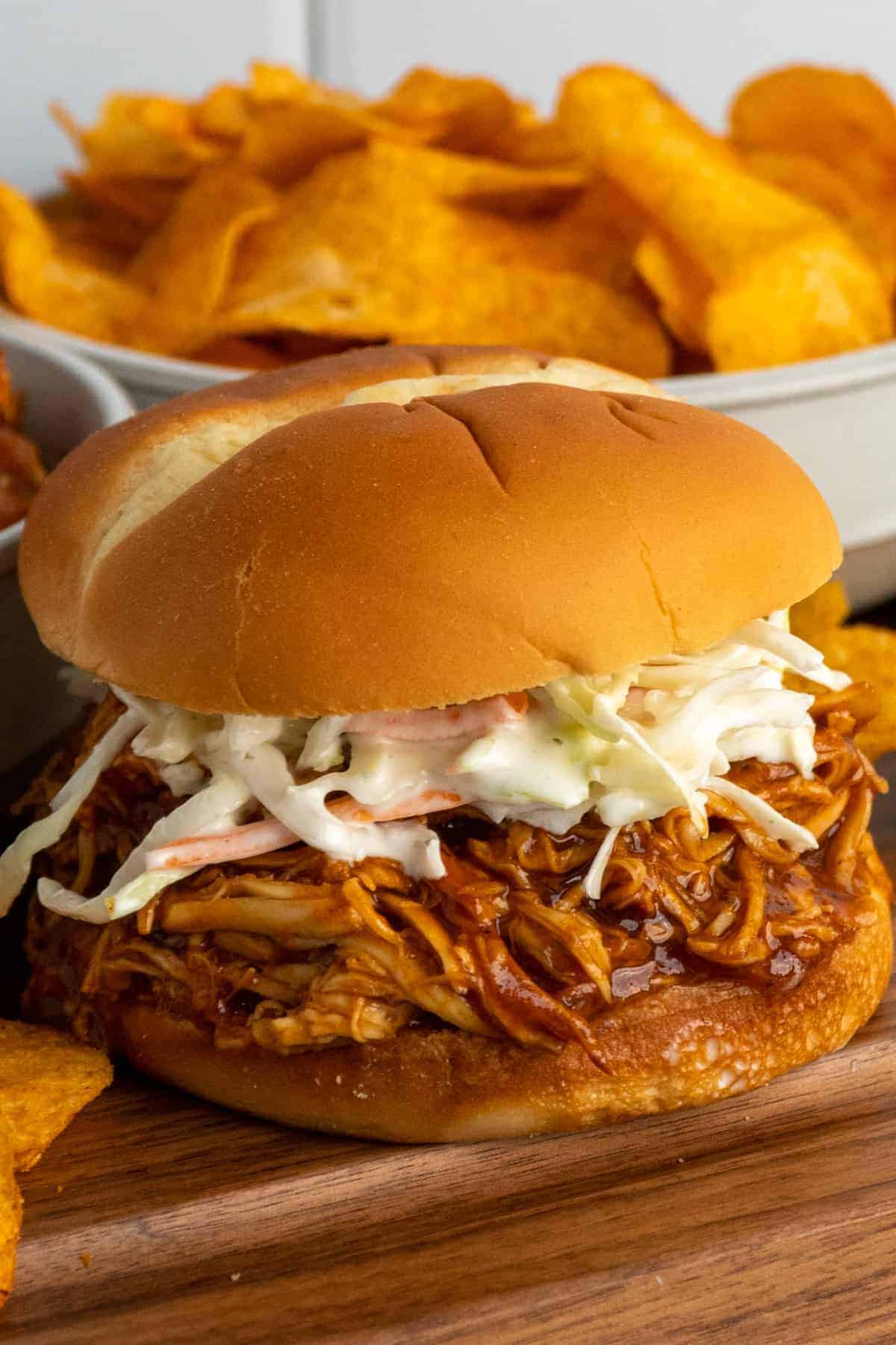 A pulled barbecue chicken sandwich topped with creamy coleslaw sits on a wooden board, with golden potato chips and a bowl of chips in the background.
