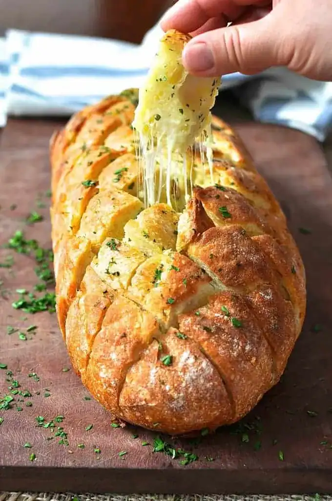 A hand pulls a cheesy, gooey piece from a loaf of garlic bread, garnished with chopped parsley, on a wooden cutting board.