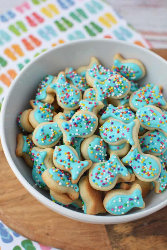 A white bowl filled with fish sugar cookies topped with blue icing and colorful sprinkles, placed on a wooden surface with a vibrant fish-patterned cloth in the background.