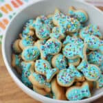 A white bowl filled with fish sugar cookies decorated with blue icing and colorful sprinkles sits on a wooden surface, next to a vibrant fish-patterned cloth.
