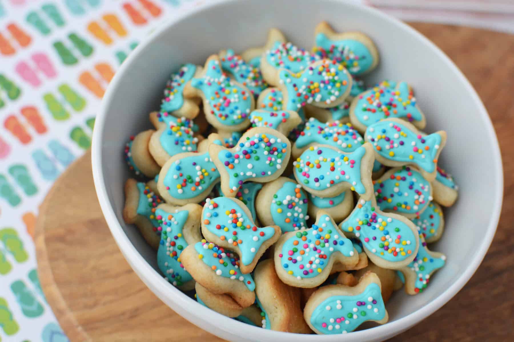 A white bowl filled with fish sugar cookies decorated with blue icing and colorful sprinkles sits on a wooden surface, next to a vibrant fish-patterned cloth.