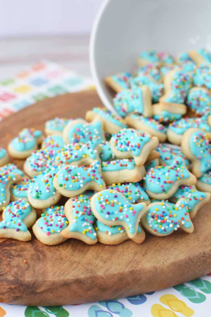 A pile of fish sugar cookies with blue icing and colorful sprinkles are scattered on a wooden board, spilling out from a tilted white bowl.