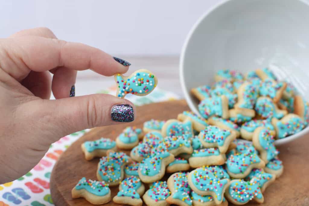A hand with glittery nail polish holds a fish sugar cookie decorated with blue icing and colorful sprinkles. More decorated cookies spill from a white bowl onto a wooden board in the background.