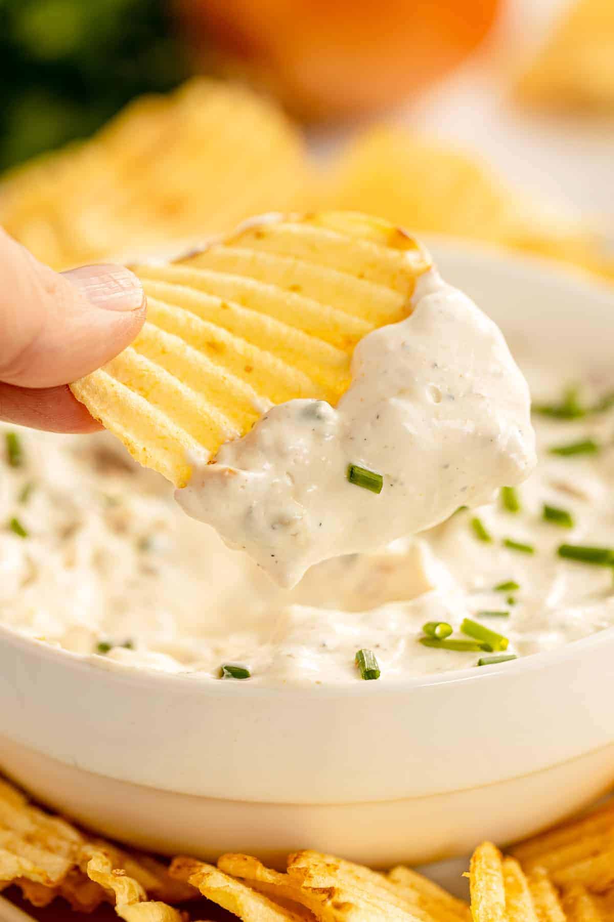 A hand holding a ridged potato chip dipped in creamy white onion dip, garnished with chopped chives, with more chips and a bowl of dip visible in the background.