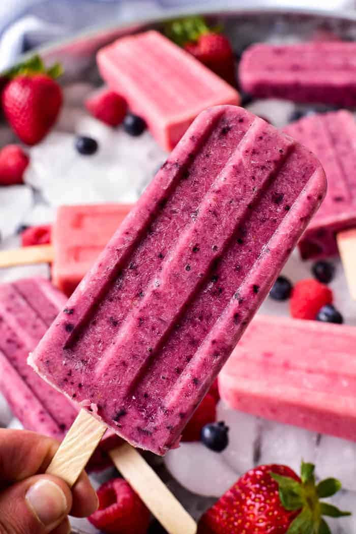 A hand holding a purple berry popsicle, surrounded by other berry popsicles, strawberries, and blueberries on a tray filled with ice.