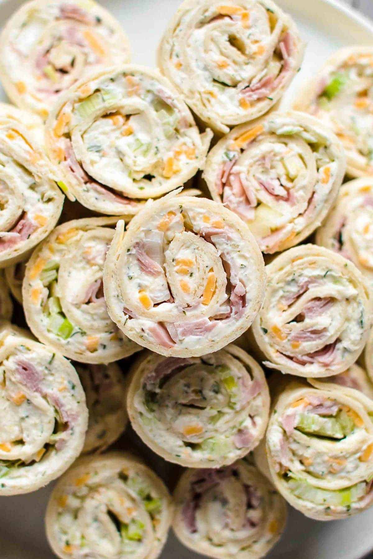 A close-up of several pinwheel sandwiches filled with cream cheese, ham, shredded cheese, and chopped green onions, arranged in a pile on a white plate.