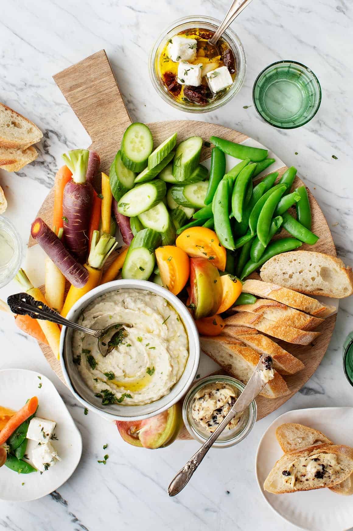 A wooden board with sliced baguette, colorful carrots, cucumber, snap peas, yellow tomatoes, and a bowl of dip. Small jars with marinated cheese and a glass of water are nearby on a marble surface.