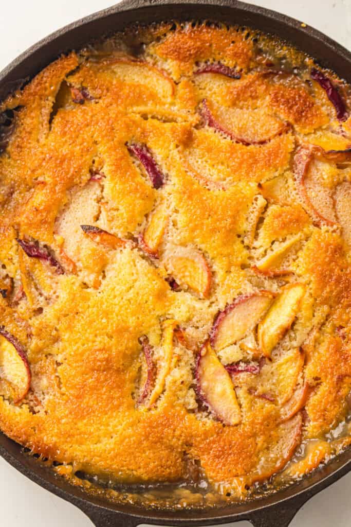 A close-up of a golden-brown peach cobbler baked in a cast iron skillet, showing juicy peach slices nestled under a crisp, caramelized biscuit topping—a mouthwatering result of this classic peach cobbler recipe.