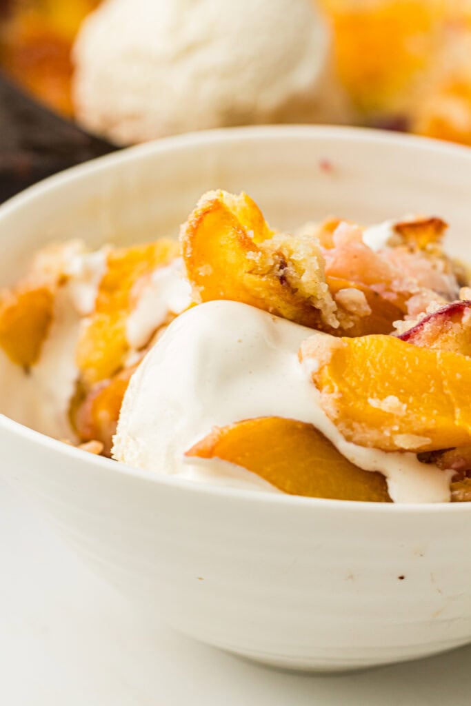 A close-up of a white bowl filled with peach cobbler and a scoop of vanilla ice cream, featuring a classic peach cobbler recipe. More dessert and ice cream are blurred in the background.
