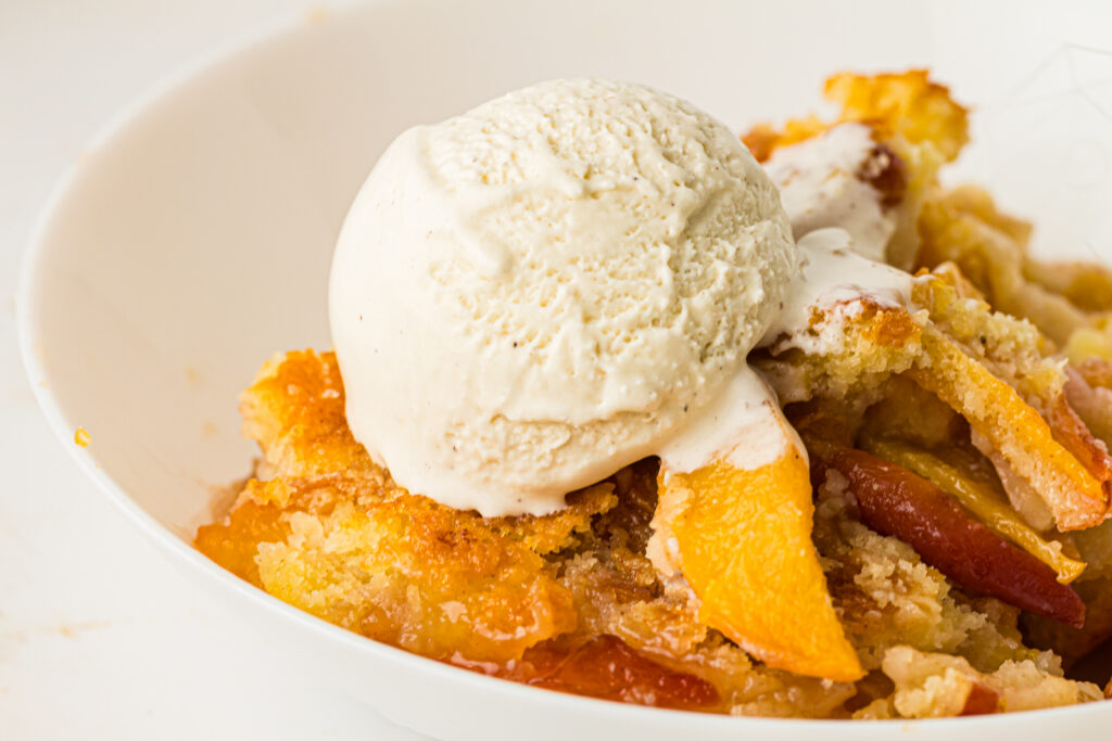 A scoop of vanilla ice cream melting on top of warm peach cobbler in a white bowl—just like the classic peach cobbler recipe you remember.