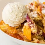 A close-up of a bowl with peach cobbler, made from a classic peach cobbler recipe and topped with vanilla ice cream, showing chunks of baked peaches and crumbly golden topping.