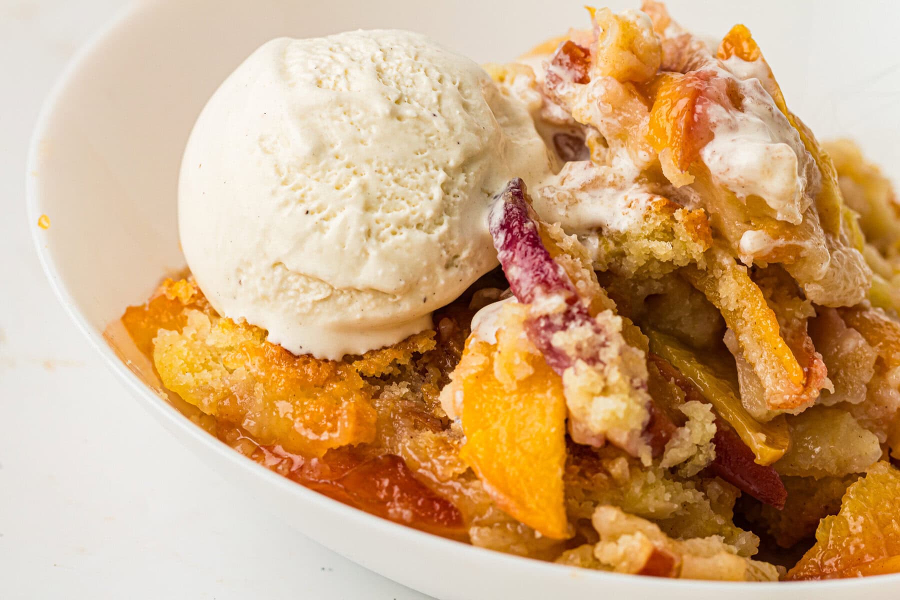 A close-up of a bowl with peach cobbler, made from a classic peach cobbler recipe and topped with vanilla ice cream, showing chunks of baked peaches and crumbly golden topping.