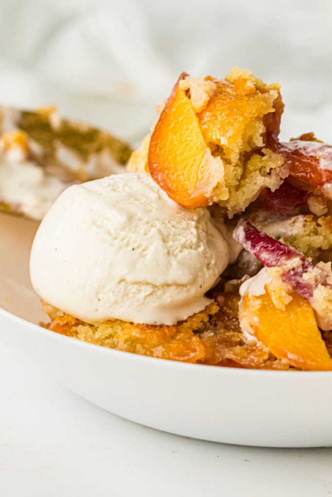 A close-up of a dessert plate with a scoop of vanilla ice cream served next to a portion of peach cobbler, featuring golden baked peaches and cake-like topping from a classic peach cobbler recipe.