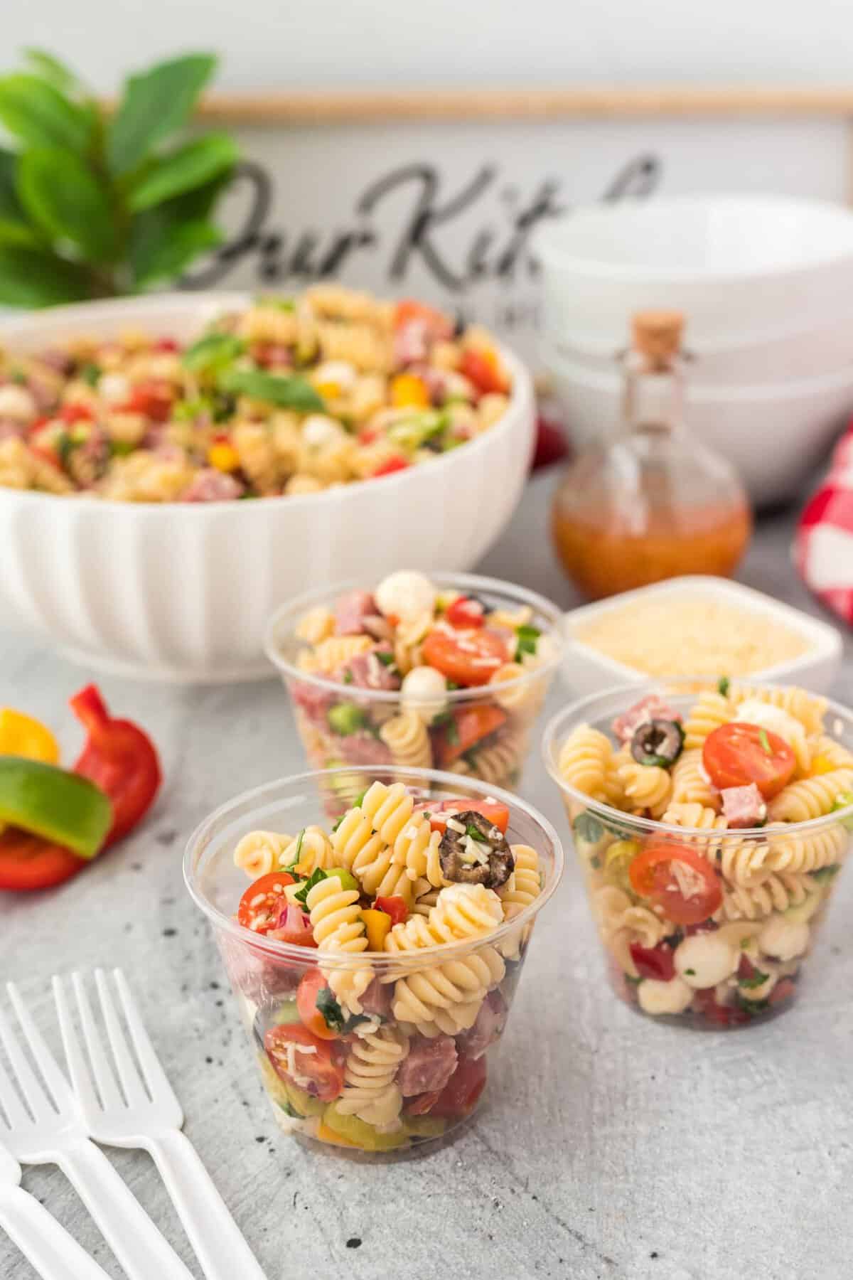 Three clear cups filled with pasta salad containing rotini, cherry tomatoes, cheese balls, and herbs. A large bowl of salad, a bottle of vinaigrette, shredded cheese, and forks are nearby on a kitchen counter.