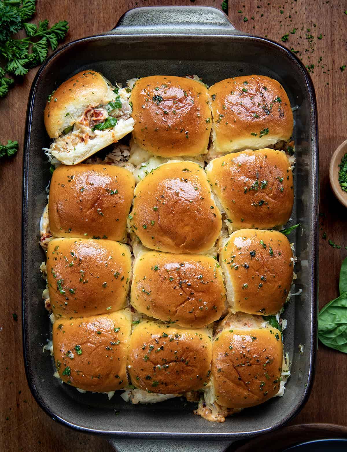 A baking dish filled with twelve golden-brown slider buns, topped with herbs, some slightly pulled apart to reveal a cheesy filling with bits of greens and tomatoes. The dish sits on a wooden surface with herbs nearby.