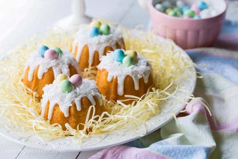 Four mini bundt cakes topped with white icing, shredded coconut, and pastel candy eggs sit on a white plate surrounded by shredded yellow paper. A pink bowl of more candy eggs is in the background, next to a pastel cloth.
