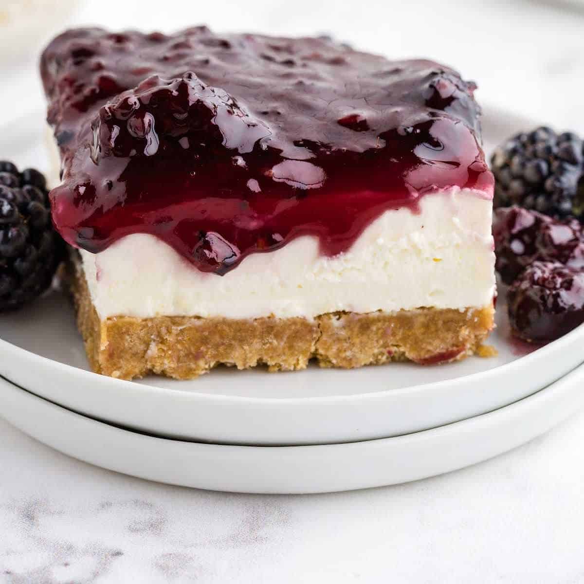 A slice of cheesecake with a graham cracker crust, creamy middle layer, and a glossy blackberry topping sits on a white plate, garnished with fresh blackberries.