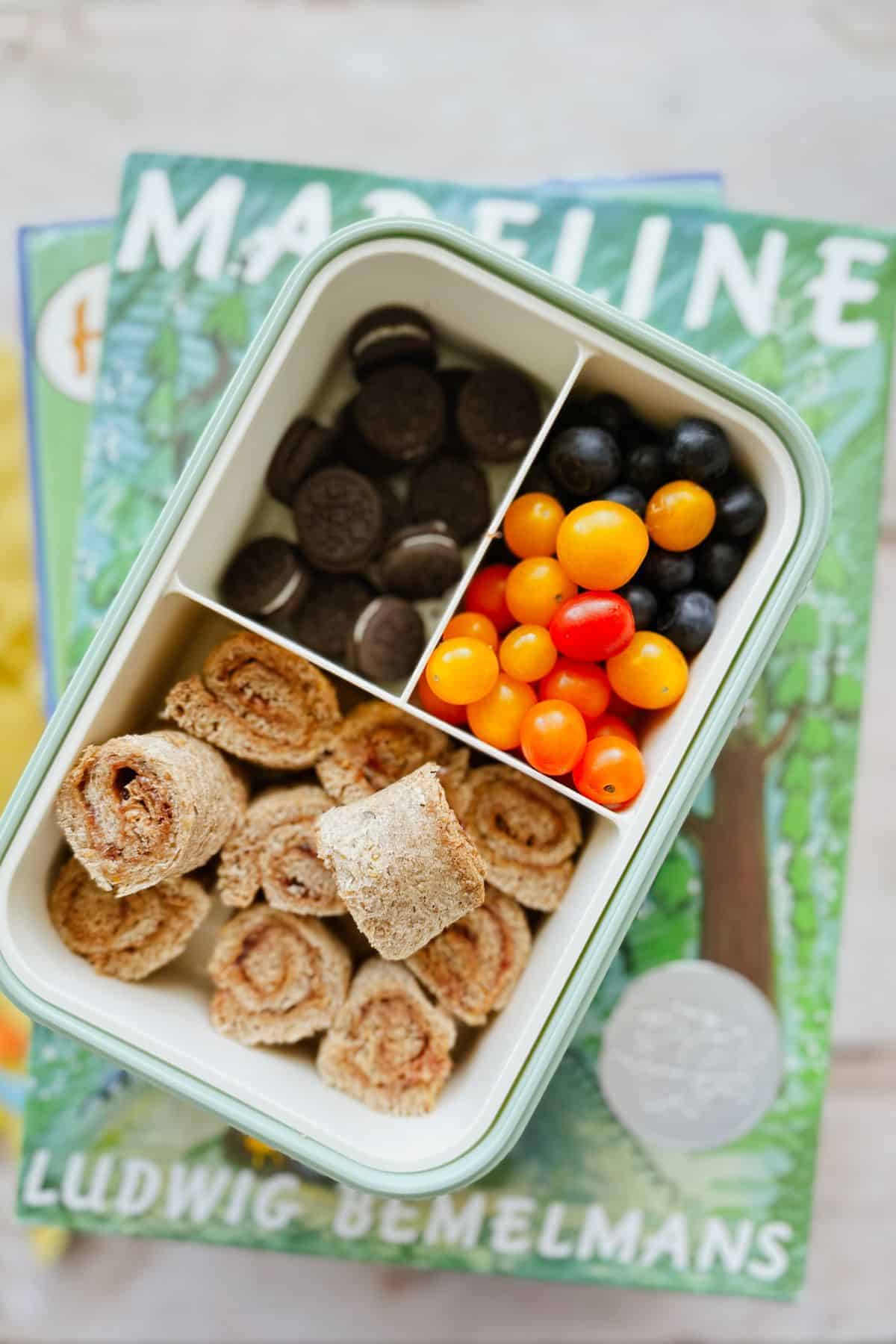 A bento box with peanut butter roll-ups, mini chocolate cookies, and a mix of yellow, orange, and black berries, placed on top of two Madeline books by Ludwig Bemelmans.