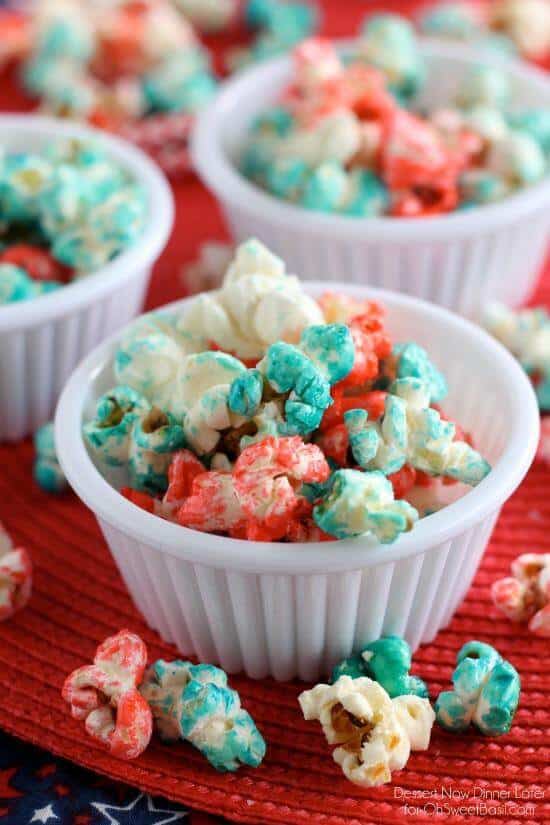 Small white cups filled with brightly colored popcorn in red, blue, and white, set on a red placemat with more cups of popcorn in the background.