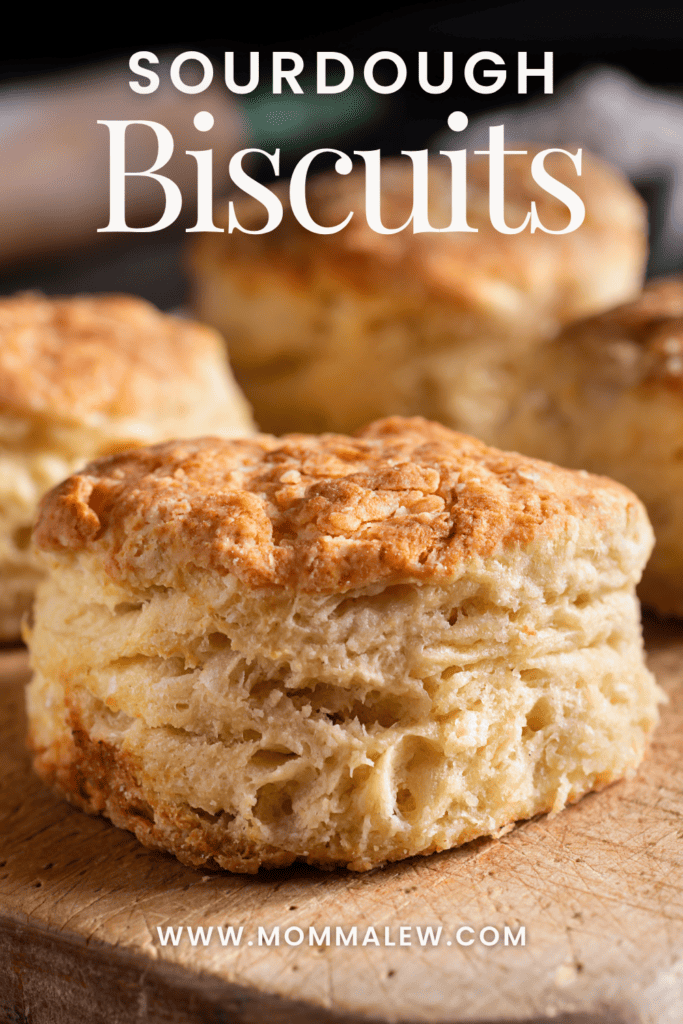 Close-up of golden brown sourdough biscuits on a wooden surface, showcasing delicious homemade sourdough biscuits. The text "Sourdough Biscuits" and www.mommalew.com appears above and below the biscuits.