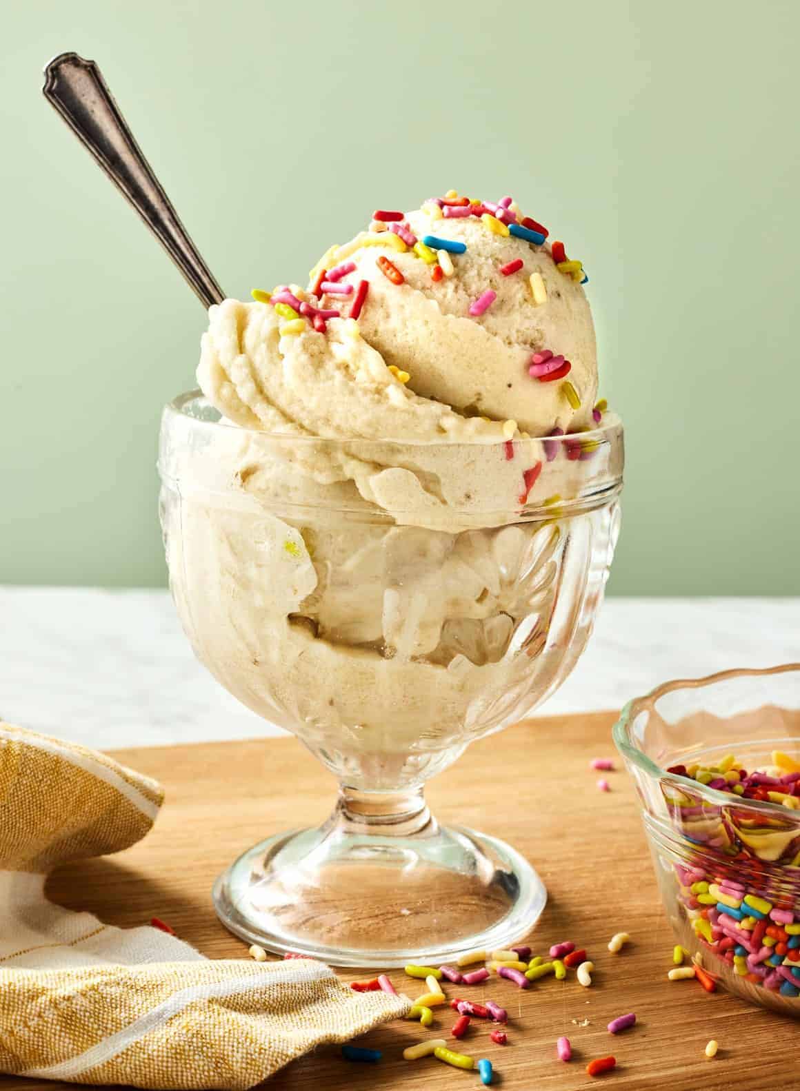 A glass dish filled with scoops of banana ice cream topped with colorful sprinkles, with a spoon inserted. A small bowl of sprinkles and a beige-striped cloth are nearby on a wooden surface.