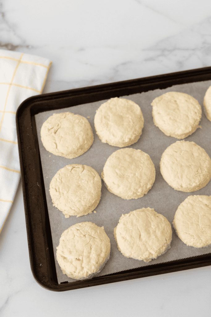 Unbaked sourdough biscuit dough rounds arranged on a parchment-lined baking sheet, ready to go in the oven. A white and yellow checkered kitchen towel is partially visible in the corner.